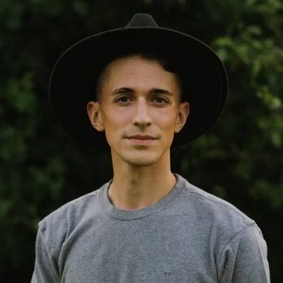 A person with short hair wearing a wide-brimmed hat and a gray T-shirt stands outdoors with a neutral expression.