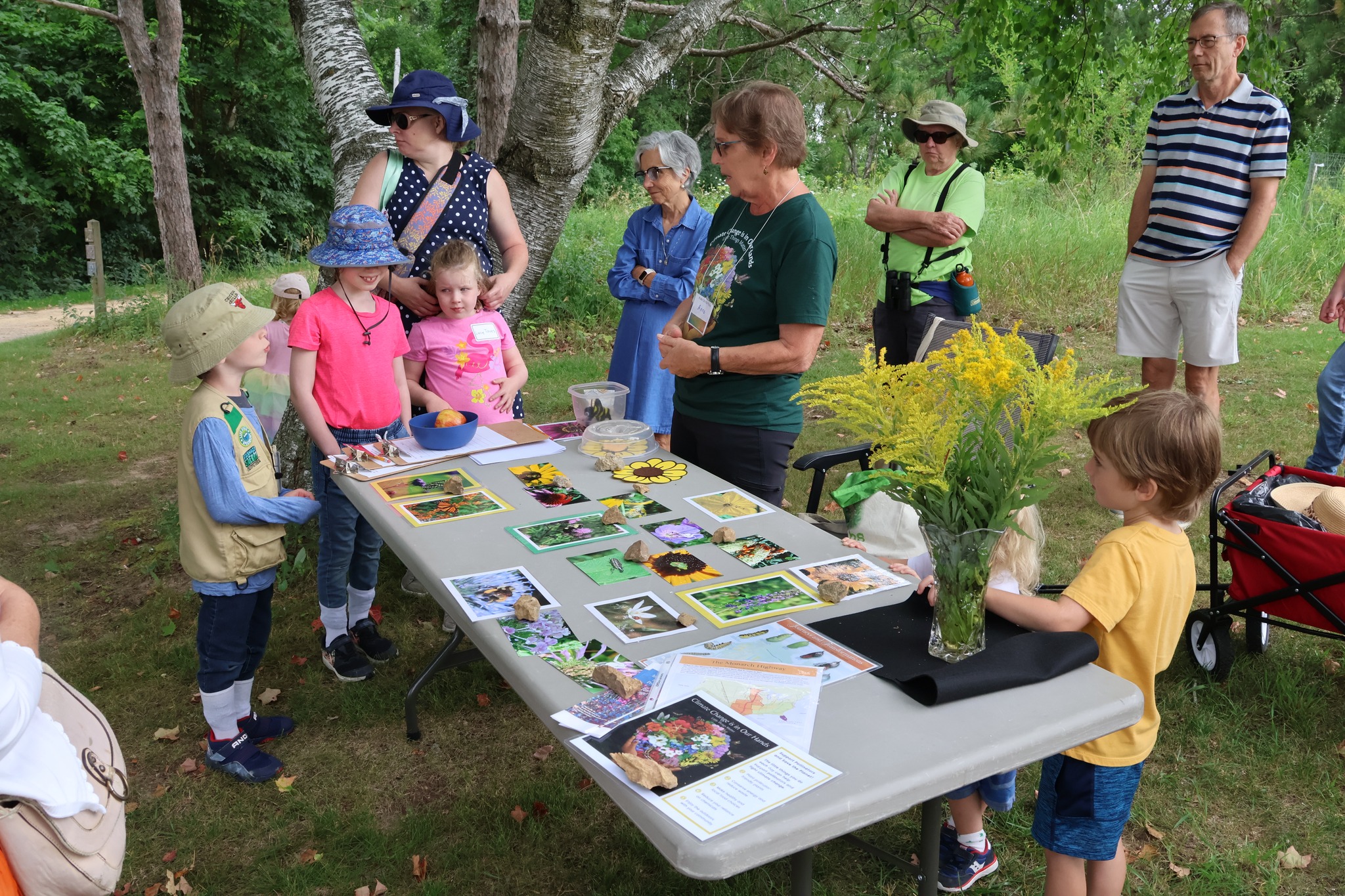 A group of children and adults gather around a table outdoors to look at various nature-themed projects and photographs. An adult is explaining the items displayed on the table.