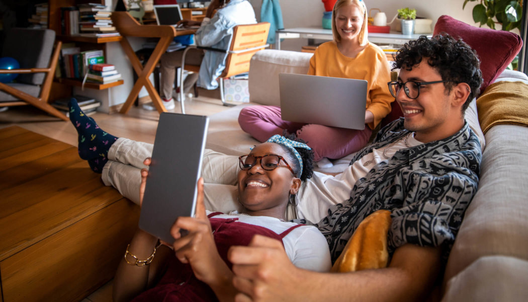 A group of friends sits on a couch watching content through OTT apps on their computer and tablet.