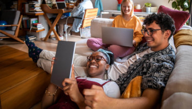 A group of friends sits on a couch watching content through OTT apps on their computer and tablet.