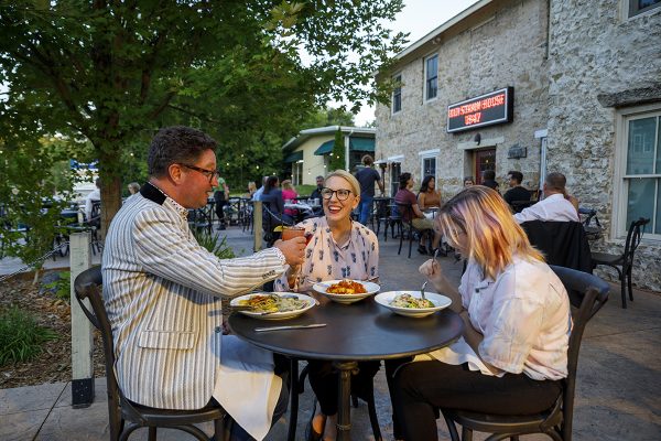 Three people dining outdoors at a restaurant, laughing and enjoying a meal together on a patio with stone buildings in the background.