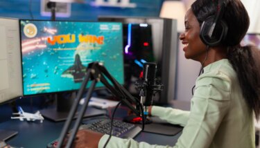 A young Black woman in a light green blouse is celebrating a victory in a video game, sitting in a professional streaming setup. She wears headphones and speaks into a microphone in front of her, with a large monitor displaying a "You Win!" screen from the game she's playing. The background shows a well-equipped gaming room with blue ambient lighting and multiple monitors, suggesting a high-tech and immersive gaming environment.