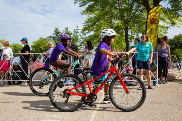 Children wearing helmets ride bicycles near a crowd of spectators. A boy in a purple shirt rides a red bike, while a girl beside him rides a turquoise bike. Trees and a yellow banner are in the background.