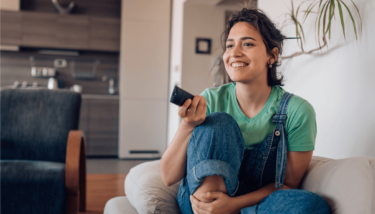 A woman sits on her couch with a remote control picking what to watch on Roku.