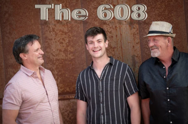 Three men stand in front of a rust-colored wall with "The 608" text above them. The man in the middle smiles directly at the camera, while the other two look at him.