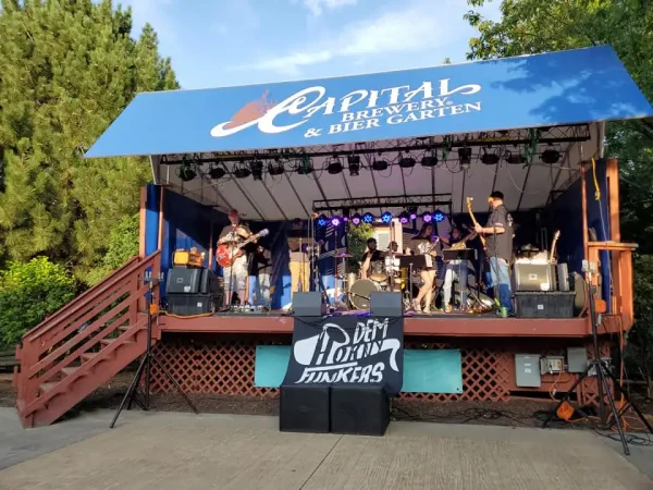 A band performs on an outdoor stage under a blue canopy that reads "Capital Brewery & Bier Garten." There are various musical instruments and equipment on the stage.