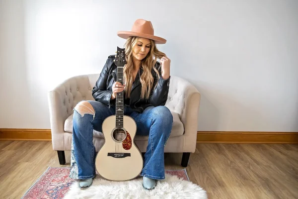 A person wearing a hat, ripped jeans, and a leather jacket sits on a couch holding an acoustic guitar.