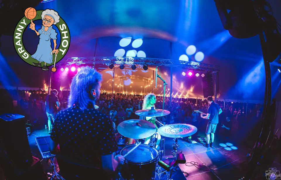 A live band performs on a brightly lit stage with a large audience. The scene includes a "Granny Shot" logo in the top left corner.