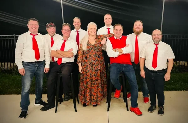 Group photo of nine people posing. Eight men are wearing white shirts and red ties, with one woman in a floral dress. Two men and the woman are seated on bar stools, while the others stand.