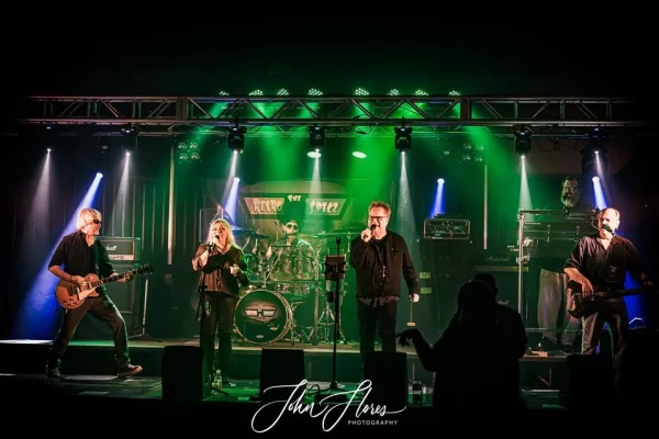 A band performs under green stage lights, featuring a guitarist, female vocalist, drummer, male vocalist, and keyboardist, with a photographer’s watermark at the bottom of the image.