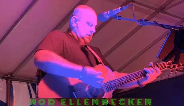 A man plays an acoustic guitar and sings into a microphone in a stage setting. Text at the bottom reads "Rod Ellenbecker.