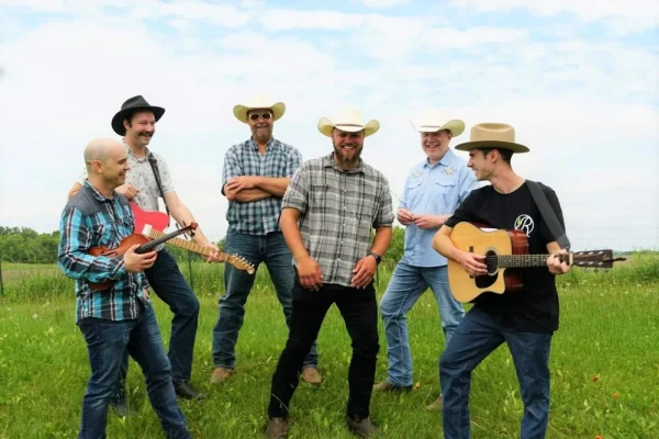 Six men wearing casual clothing and cowboy hats stand in a grassy field, playing musical instruments and smiling.