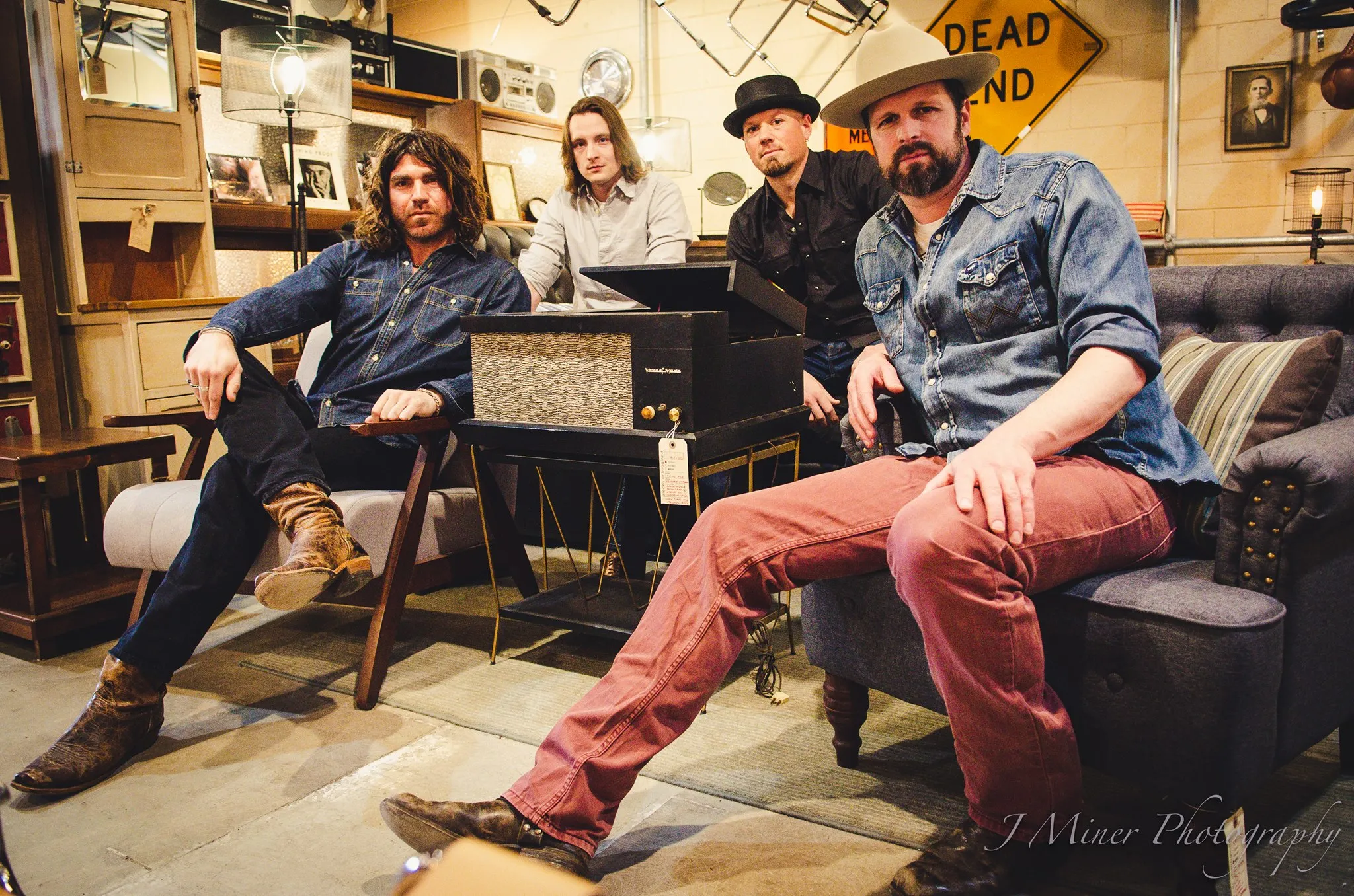 Four men, casually dressed, sit and stand around vintage audio equipment in a room with various retro decorations. One wears a cowboy hat and another wears a fedora.