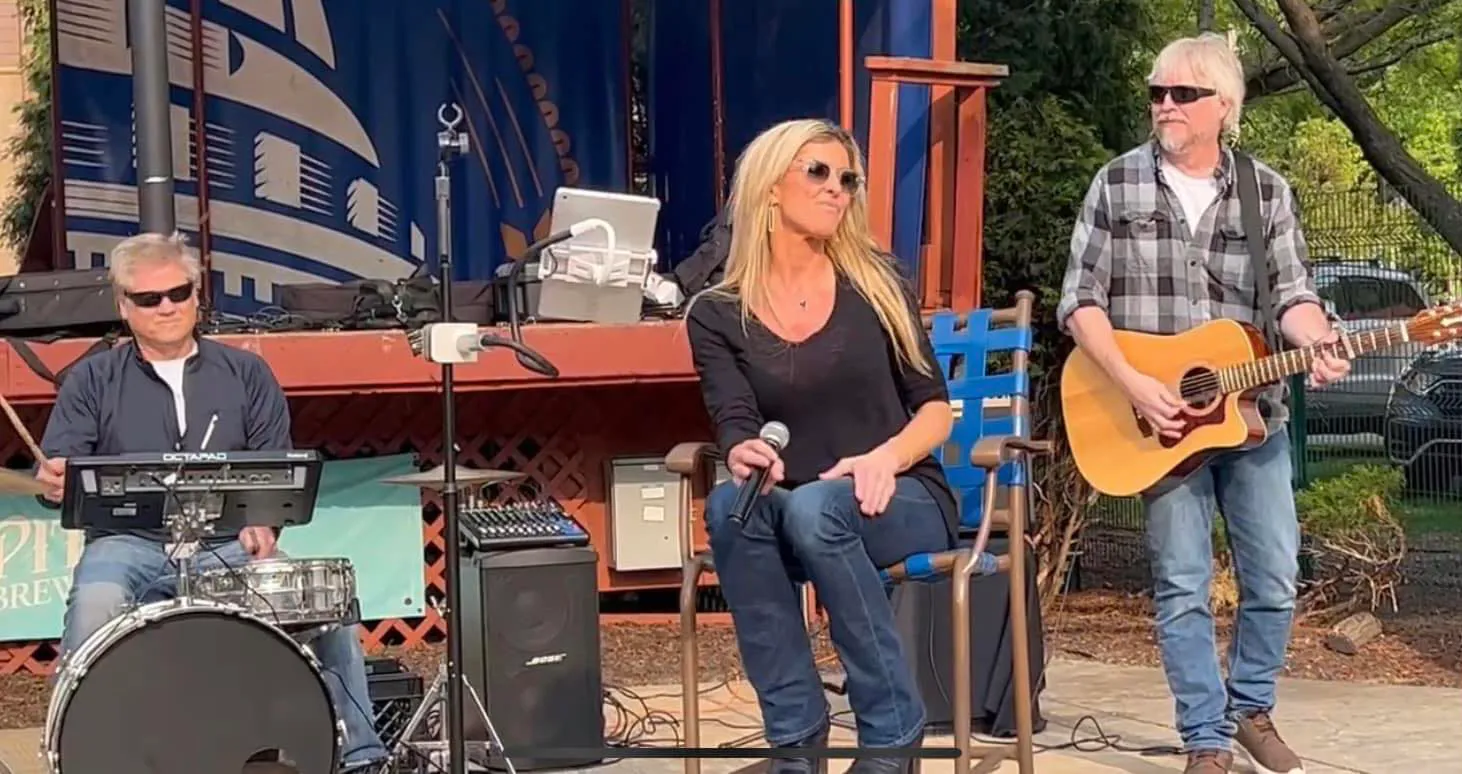 Three musicians perform outdoors. A seated woman holding a microphone is in the center, a guitarist is standing on the right, and a keyboard player is on the left.